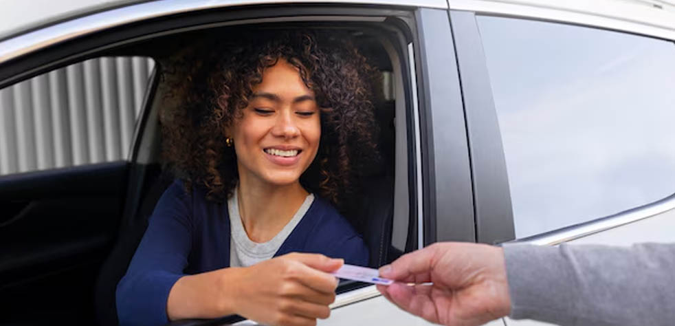 Une personne assise au volant d'une voiture sourit en tendant son permis de conduire à une autre personne située à l'extérieur du véhicule.